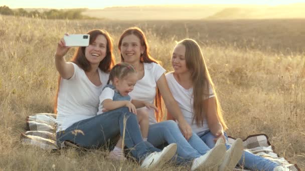 Šťastná rodina cestuje, selfie na smartphonu v parku. Máma a zdravé děti sedí na dece. matka se svými dcerami je fotografována na poli na přikrývce. rodinné cestování a dobrodružství - Záběry, video