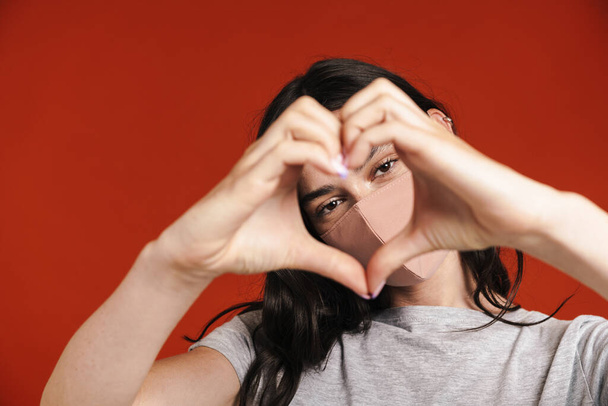 Image of young woman in face mask making heart gesture with hands isolated over red background - Fotografie, Obrázek