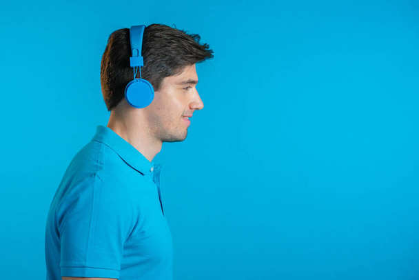 Handsome young man with trendy hairdo having fun, smiling with headphones in studio against blue background. Music, dance, radio concept. - Φωτογραφία, εικόνα