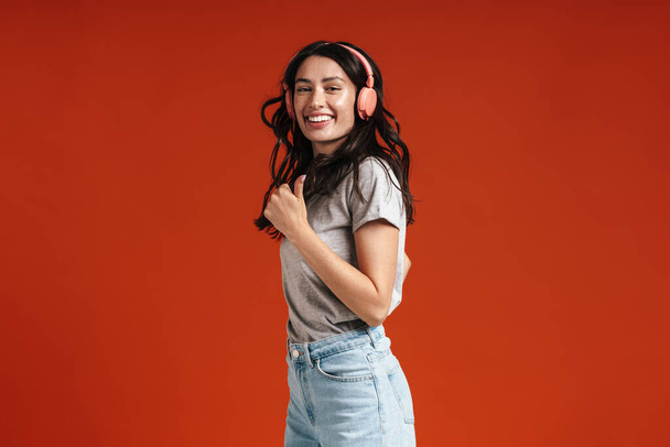 Image of young happy woman showing thumb up and using headphones isolated over red background - Foto, immagini