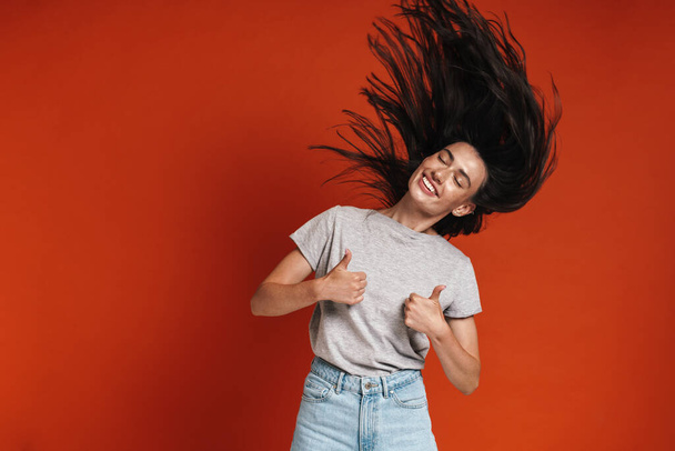 Image of beautiful happy woman showing thumbs up and shaking her hair isolated over red background - Фото, зображення