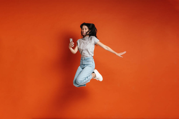 Image of young joyful woman jumping and using cellphone isolated over red background - Zdjęcie, obraz