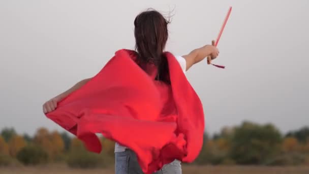 child play knights. free girl in red cloak runs with swords in his hand across field playing medieval knight. young girl playing super heroes. children fight with toy sword. happy childhood - Footage, Video
