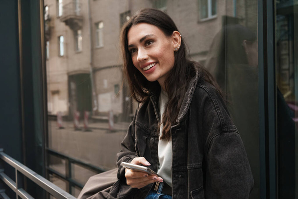 Photo of beautiful cheerful happy young woman walking outdoors by the street and using mobile phone - Фото, зображення