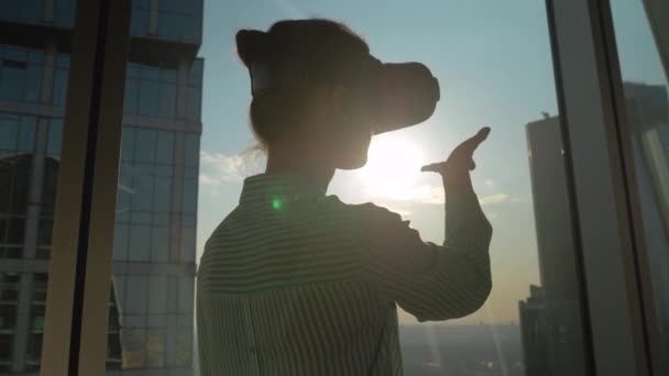 Zwolniony ruch: kobieta używająca zestawu słuchawkowego VR przeciwko oknie drapacza chmur w biurze - Materiał filmowy, wideo
