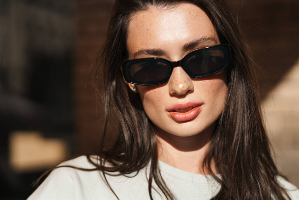 Image of beautiful serious young woman posing outdoors wearing sunglasses - Фото, изображение