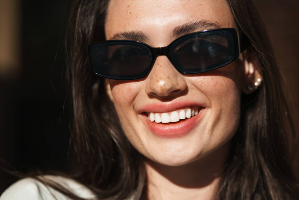 Image of beautiful happy optimistic young woman posing outdoors wearing sunglasses - Foto, afbeelding