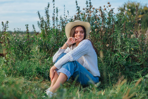 Young beautiful brunette woman dressed in a white sweater, jeans and cowboy straw hat sitting on green grass, smiling and laughing during sunset - Fotoğraf, Görsel