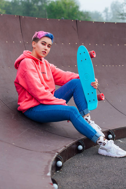 Stylish young woman with short colored hair sitting with her blue plastic skateboard in skatepark. Youth concept. - Fotó, kép