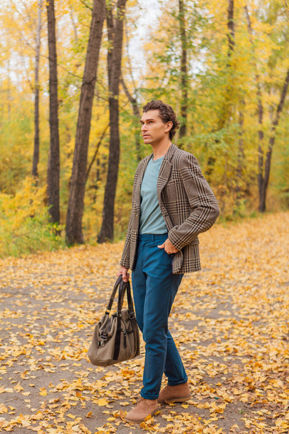 Tall handsome man dressed in a brown jacket walking with a bag on the autumn alley - Valokuva, kuva