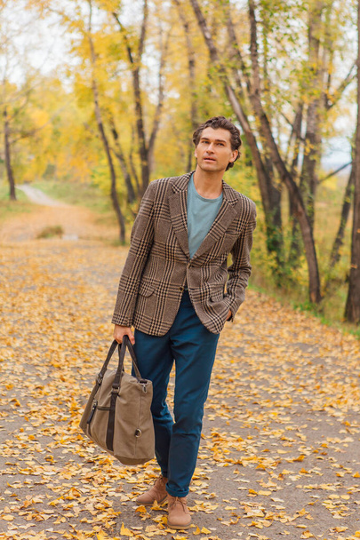 Tall handsome man dressed in a brown jacket walking with a bag on the autumn alley - Fotografie, Obrázek