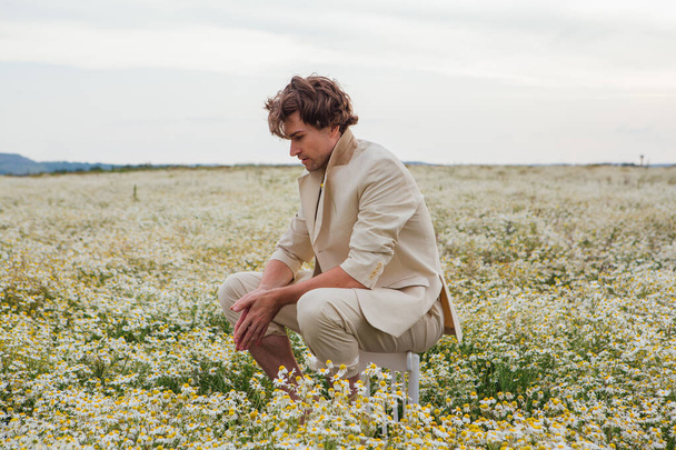 Tall handsome man dressed in a white suit on naked body sitting on a back of a chair in camomile flowers field - Fotoğraf, Görsel
