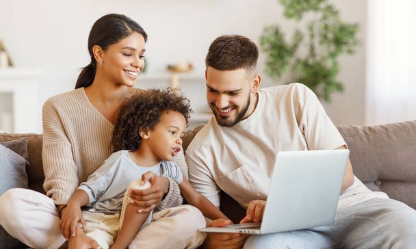 Счастливые многоэтнические родители и удивленный мальчик смешанной расы смотрят видео на ноутбуке, отдыхая дома на диване - Фото, изображение