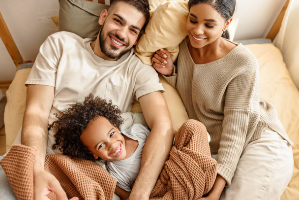 幸せな家族の多民族の母、父と息子は笑い、遊び、そして家のベッドルームでベッドでタックル - 写真・画像