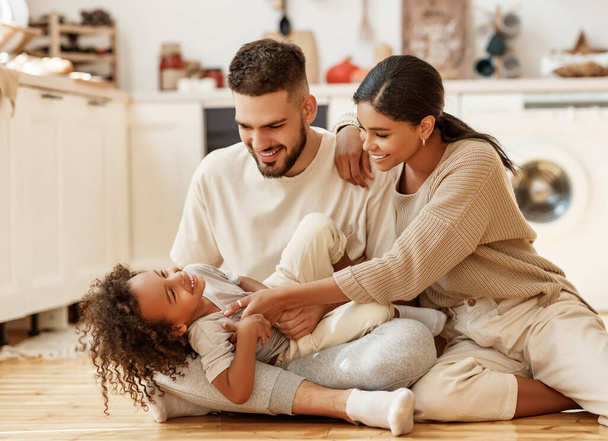 familia feliz madre multiétnica, padre e hijo riendo, jugando, y cosquillas en el suelo en la acogedora cocina en hom - Foto, Imagen