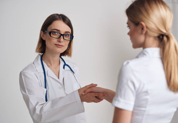 Nainen lääkäri lääketieteellisessä kaapussa kättelee potilasta valkoisessa t-paidassa kevyellä taustalla - Valokuva, kuva