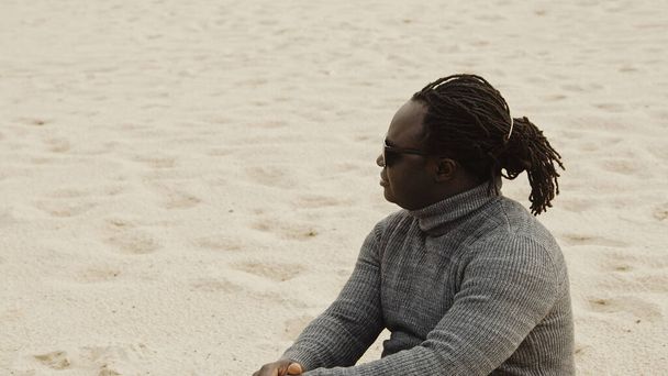 Νεαρός Αφροαμερικάνος μαύρος κάθεται στην άμμο με πουλόβερ και χαλαρώνει στις χειμερινές διακοπές - Φωτογραφία, εικόνα