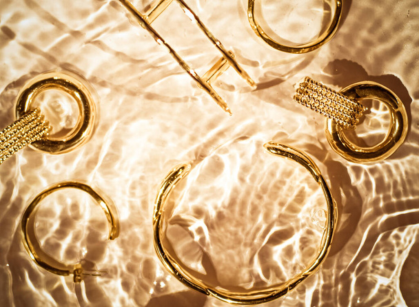 Gouden armbanden, oorbellen, ringen, sieraden op goud water achtergrond, luxe glamour en vakantie schoonheid ontwerp voor sieraden merk advertenties - Foto, afbeelding