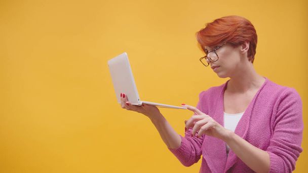 Zdezorientowana ruda kobieta w średnim wieku trzymająca laptopa i kartę kredytową. Koncepcja zakupów online. izolowany na pomarańczowym tle - Zdjęcie, obraz