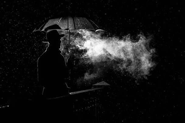 silueta de un hombre con sombrero bajo un paraguas en la noche bajo la lluvia en la ciudad - Foto, imagen