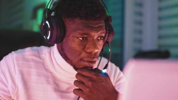Homem preto jovem com fone de ouvido iluminado usando laptop. Home office e conceito de trabalho remoto - Filmagem, Vídeo