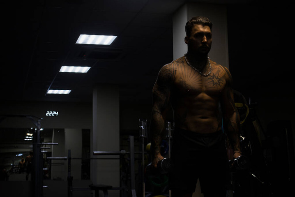 Lihaksikas uros urheilija, jolla on tatuoitu alaston vartalo seisomassa pimeässä kuntosalilla ja tekemässä käsipainokiharoita harjoittelun aikana - Valokuva, kuva
