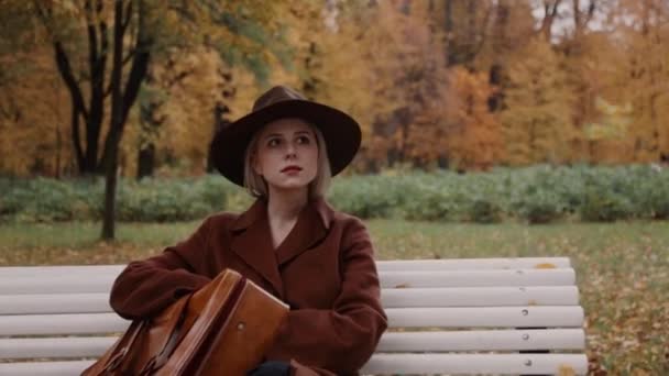 Chica de estilo en abrigo marrón con maleta sentada en un banco en el parque de otoño - Imágenes, Vídeo