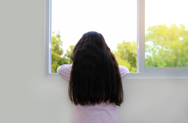 Vista trasera caucásica niña en casa se encuentra en la ventana y mirando hacia fuera. - Foto, Imagen