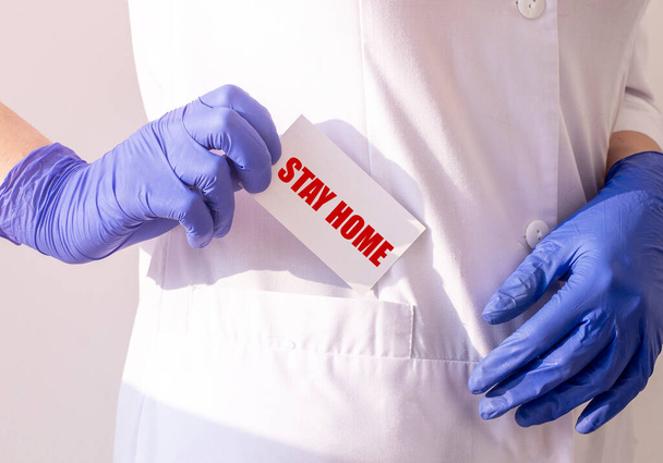 Γιατρός με μπλε ιατρικά γάντια κρατά ένα λευκό φύλλο με την επιγραφή μείνετε στο σπίτι. Κλείσε.. - Φωτογραφία, εικόνα