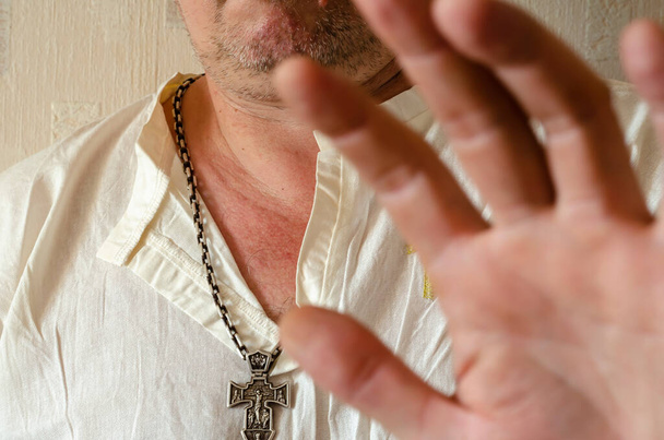 Мужчина средних лет в белой льняной рубашке с христианским крестом на шее закрывает руку от камеры. Небритый мужчина с массивной серебряной цепью и крестом. Фокус на заднем плане - Фото, изображение