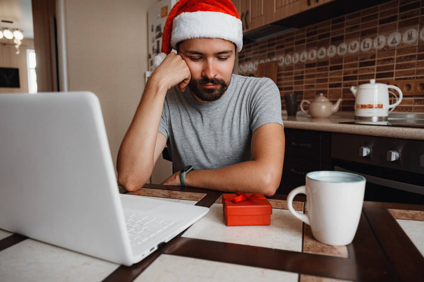 Muž padající v depresi a osamělý během Vánoc. Virtuální vánoční večírek. Online tým setkání videokonference volání z domova. Muž v Santově klobouku - Fotografie, Obrázek