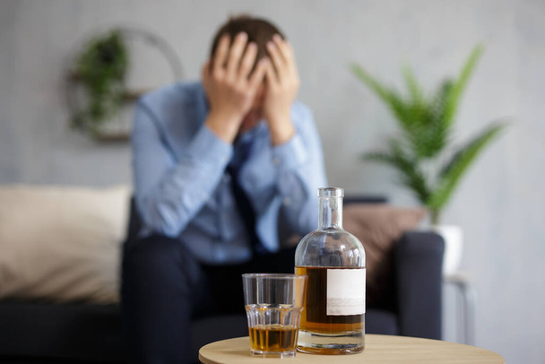 zblízka láhev whisky a sklo, depresivní a unavený podnikatel s bolestí hlavy sedí doma nebo v kanceláři - Fotografie, Obrázek