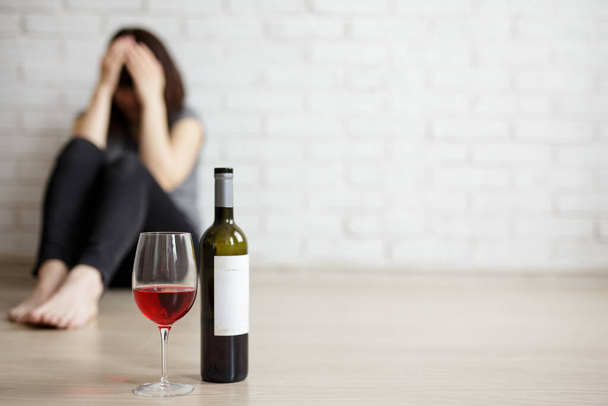 alcoolismo feminino, relacionamento, divórcio e depressão conceito - close-up de vidro e garrafa de vinho e mulher estressada chorando e sentado no chão, espaço de cópia sobre fundo da parede de tijolo branco - Foto, Imagem