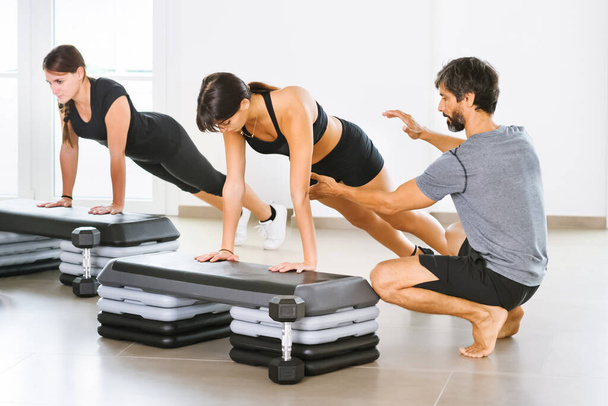 Entrenador personal masculino joven que ayuda a una mujer que hace una pose de tabla de yoga en una clase en un gimnasio aireado brillante en un concepto de salud y fitness o estilo de vida saludable - Foto, Imagen