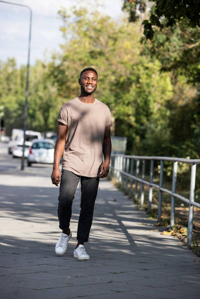Молодий чорношкірий чоловік, що йде міським тротуаром, посміхається. Повна довжина. Вертикальний
.  - Фото, зображення