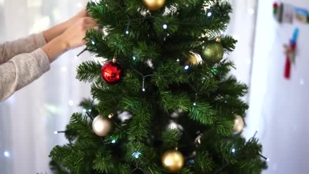 Una mujer joven decora con bolas de Navidad un esponjoso árbol de Navidad en la habitación. Hermoso fondo brillante de Navidad. humor navideño festivo. - Imágenes, Vídeo
