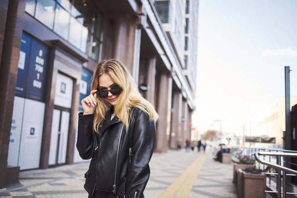 Fashionable woman in a leather jacket and sunglasses near the building  - Фото, зображення