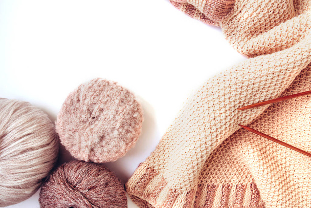 Balls of woolen yarn of nude colors and a knitted beige sweater, knitting needles on a white background. Handmade woolen clothes. Traditional hobby. Copy space - Photo, Image