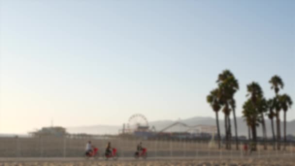 California beach aesthetic, people ride cycles on a bicycle path. Blurred, defocused background. Amusement park on pier and palms in Santa Monica american pacific ocean resort, Los Angeles CA USA - Footage, Video
