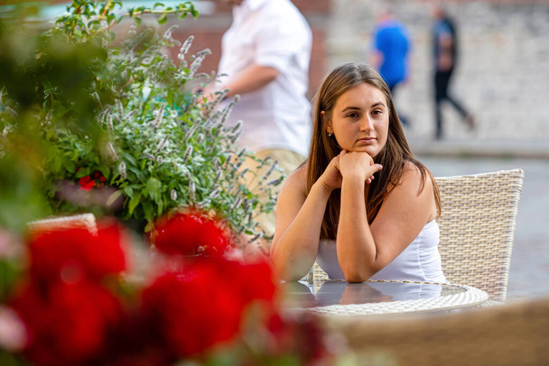  young beautiful woman sitting at table of street cafe and waiting for meeting, defocused flowers in the foreground - Zdjęcie, obraz