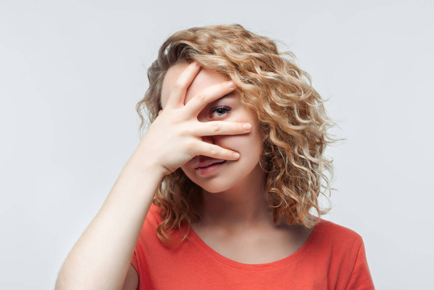 Retrato de jovem assustada com cabelos encaracolados em camiseta casual cobrindo os olhos com as mãos. Emoções humanas, conceito de expressão facial. Estúdio tiro, fundo branco, isolado - Foto, Imagem
