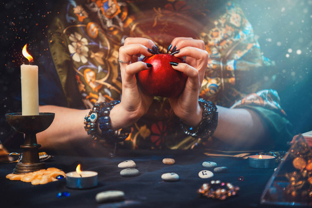 Astroloji ve kehanet. Bir falcı elinde kırmızı bir elma tutuyor. Masanın üzerinde rünler, mumlar, tılsımlar vardı. Ellerin yakın çekimi. - Fotoğraf, Görsel