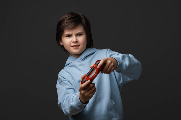Aranyos fiú portréja 10-12 éves, alkalmi ruhákba öltözve, joystickkal a kezében áll és a kamerába néz. Gyerekkor, arckifejezés. Stúdió lövés, szürke háttér - Fotó, kép