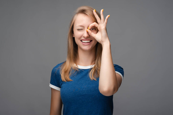 金髪のストレートの髪で陽気な笑顔の女性は、白い歯を実証し、 OKAYジェスチャーで指を介してカメラを見て。顔の表情感情の概念. - 写真・画像