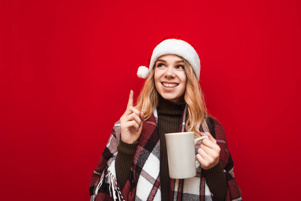Happy girl in plaid and christmas hat stands on red background with cup of hot drink in her hand and has ideas, looks away and smiles, lifted finger up. Christmas portrait of a cute girl with a cup - Photo, Image