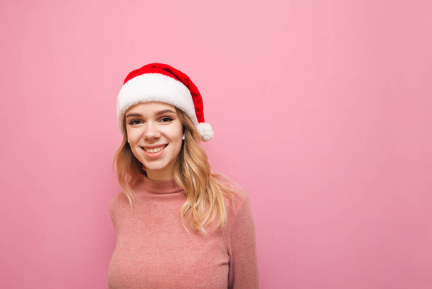 Happy girl in cordless headphones stands on a pink background in a Christmas hat, looks into the camera with a smile on her face. Positive girl listens to music in wireless headphones and laughs. - Foto, afbeelding