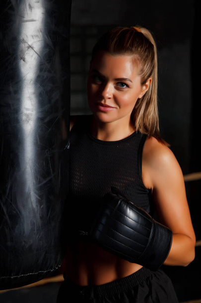 Pretty young female boxer is standing in ring at punching bag in an old gym, her hands in Boxing gloves. Woman in box training. Concept of healthy lifestyle, sports and exercise in gym. Copy space - Zdjęcie, obraz