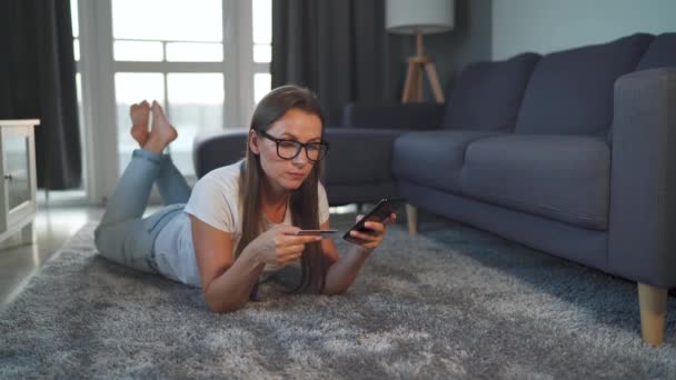 Gözlüklü mutlu kadın yerde yatıyor ve kredi kartı ve akıllı telefon kullanarak online alışveriş yapıyor. İnternetten alışveriş, yaşam tarzı teknolojisi. - Video, Çekim