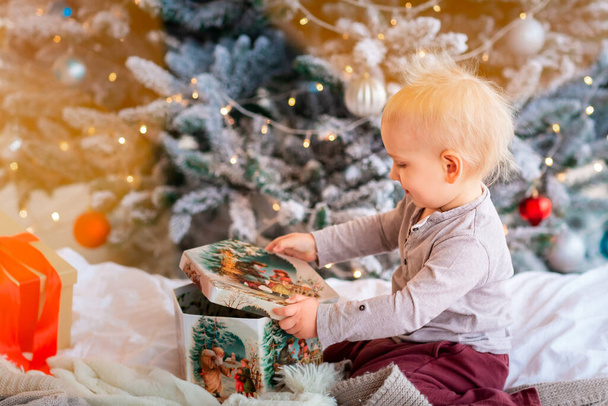 Happy little baby boy opening present boxes near Christmas tree. Christmas morning concept - Photo, Image
