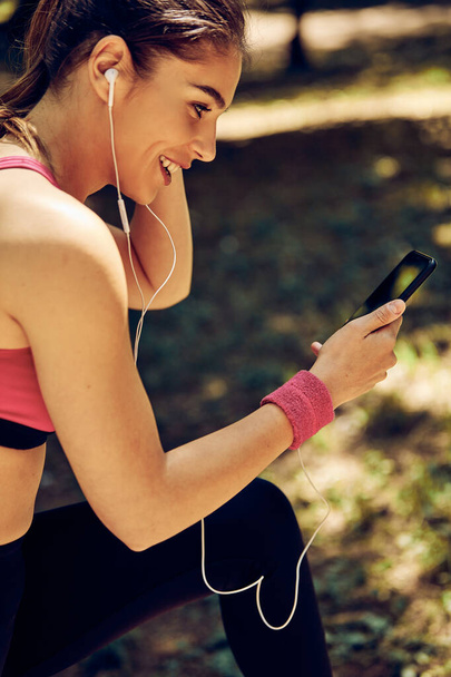 Smiling attractive fit sportswoman using phone and taking a break after running in nature. - Photo, Image